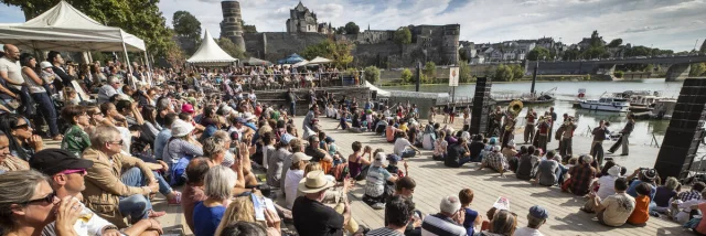 Foule regroupée au bord de la Maine, écoutant un orchestre jouer. Journée ensoleillée d'été.