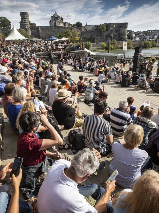 Foule regroupée au bord de la Maine, écoutant un orchestre jouer. Journée ensoleillée d'été.