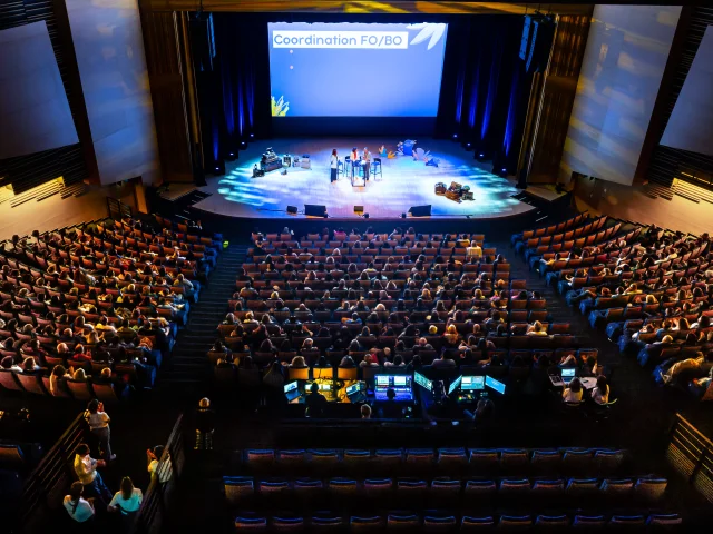 Plan large en plongée du grand auditorium dans le noir, seule la scène est éclairée