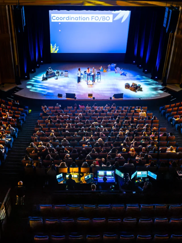 Plan large en plongée du grand auditorium dans le noir, seule la scène est éclairée