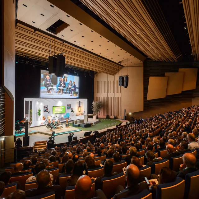 Plan large du grand auditorium avec public sur les fauteuils et présentateurs sur scène