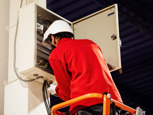 Homme en veste rouge avec un casque blanc regarde dans un tableau électrique