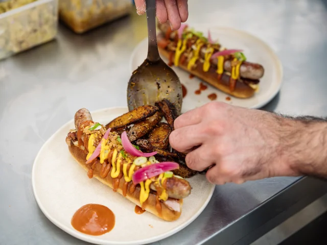 Hot dog maison préparé par le chef, gros plan sur ses mains qui dressent l'assiette.