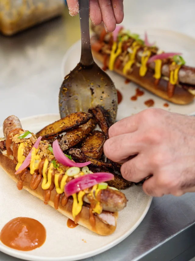 Hot dog maison préparé par le chef, gros plan sur ses mains qui dressent l'assiette.