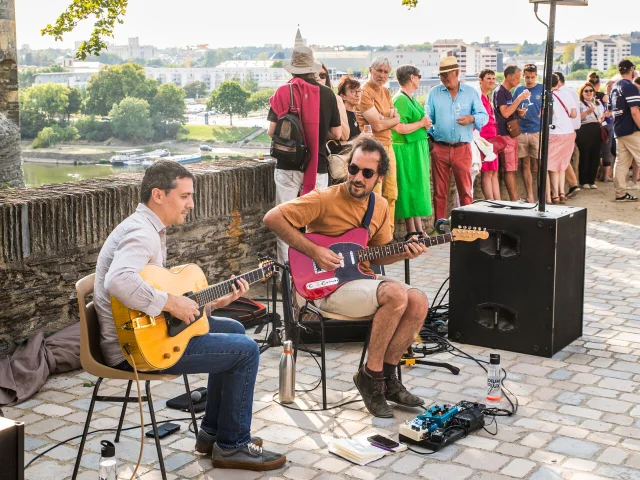 Musiciens jouant assis de la guitare sous un arbre à l'ombre devant quelques spectateurs