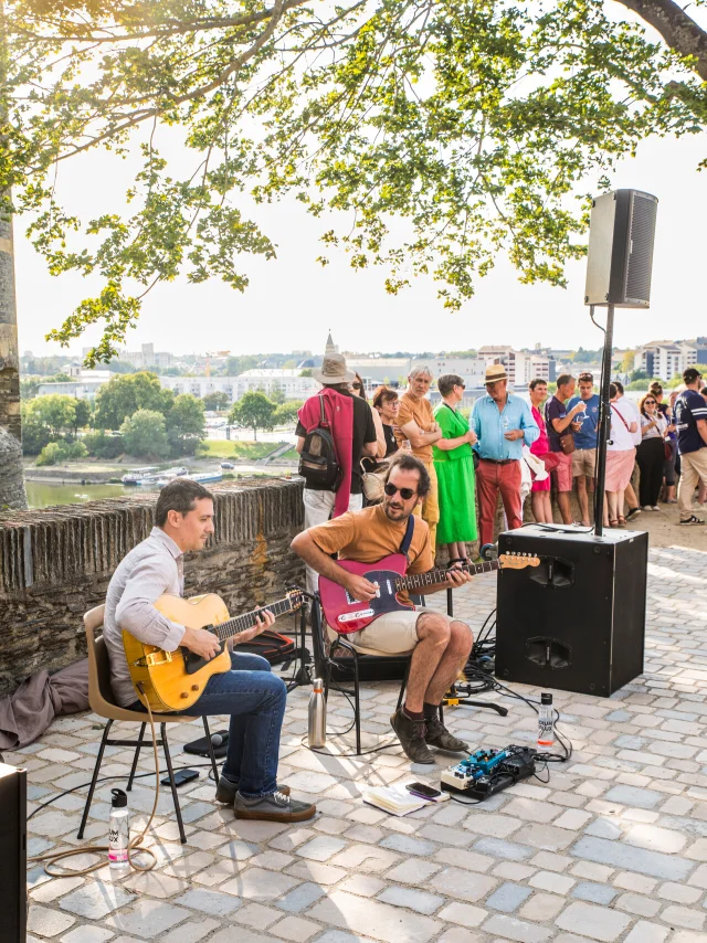 Musiciens jouant assis de la guitare sous un arbre à l'ombre devant quelques spectateurs