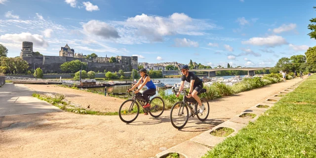 Une femme et un homme sur des vélos gravel passent au bord du port d'Angers, cale de la Savatte, Château et rivière en arrière plan