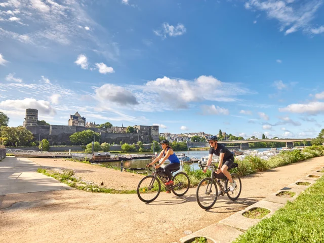 Une femme et un homme sur des vélos gravel passent au bord du port d'Angers, cale de la Savatte, Château et rivière en arrière plan