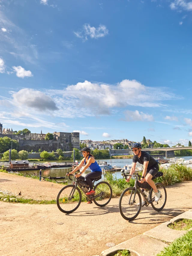 Une femme et un homme sur des vélos gravel passent au bord du port d'Angers, cale de la Savatte, Château et rivière en arrière plan