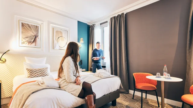 Un couple dans une chambre d'hôtel, lui à la fenêtre consulte un plan, elle, assise sur le lit, le regarde