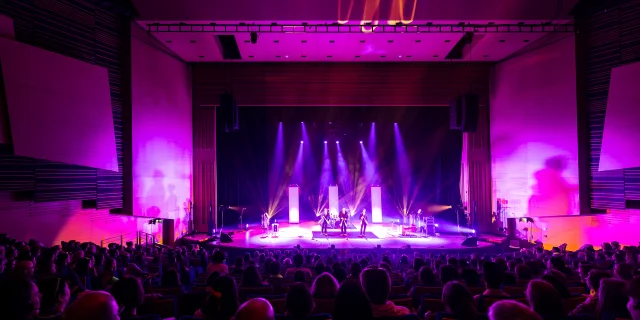 Plan large de la scène du grand auditorium du Centre de congrès éclairée dans les tons violet rose, la salle est comble.